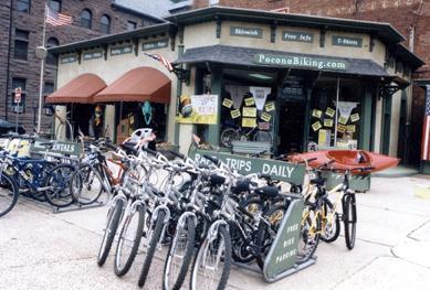 A storefront for Pocono Biking featuring bicycles parked in front and promotional signs, with a green and yellow color scheme. The building has awnings and large windows, displaying various biking-related advertisements and information.
