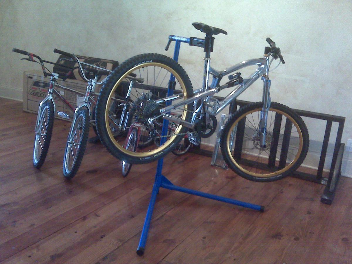 A shiny silver mountain bike mounted on a blue bike repair stand, with two other bicycles in the background. The scene is set indoors on wooden flooring with a neutral wall. The bikes vary in style, showcasing the distinct features of each.