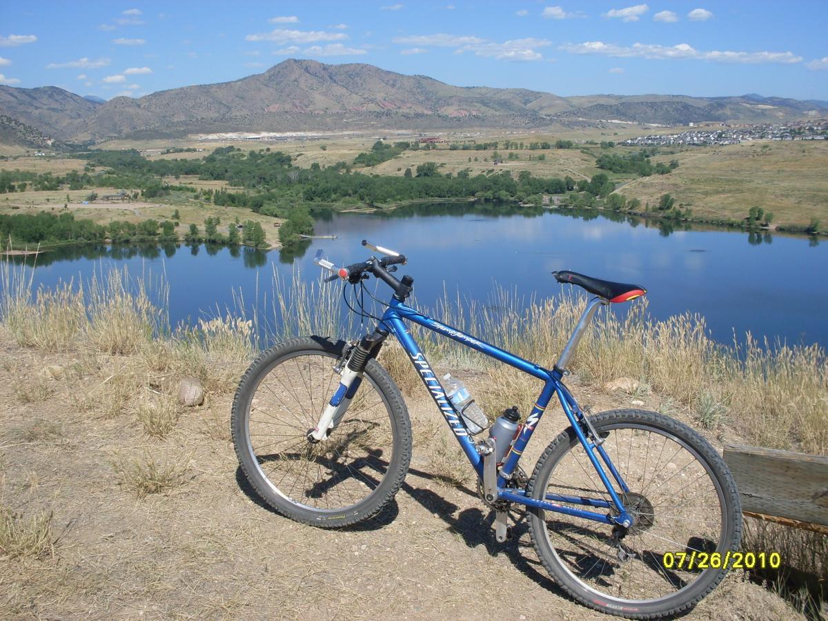 A mountain bike parked on a hilltop overlooking a serene lake, surrounded by lush greenery and distant mountains under a clear blue sky. Bear Creek Lake Park mountain bike trail.