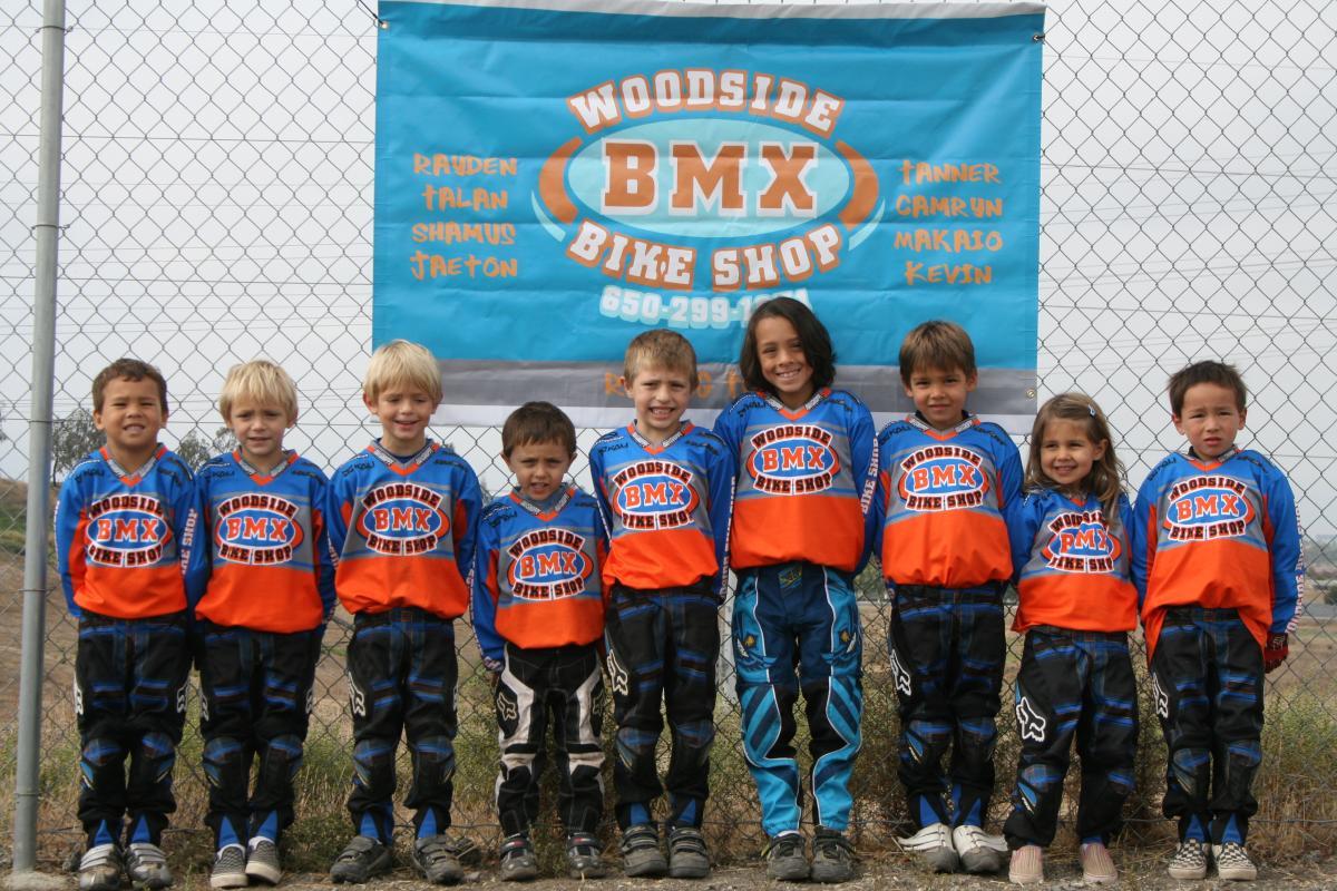 A group of nine children, dressed in matching BMX jerseys and pants, stand in front of a blue banner for Woodside BMX Bike Shop. They are smiling and posing together, showcasing a sense of camaraderie and enthusiasm for BMX biking. The background features a chain-link fence and grassy terrain.
