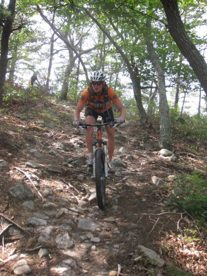 A mountain biker navigating a rocky trail through a wooded area, wearing a helmet and an orange jersey, with sunlight filtering through the trees. North Fork Mountain Trail mountain bike trail.