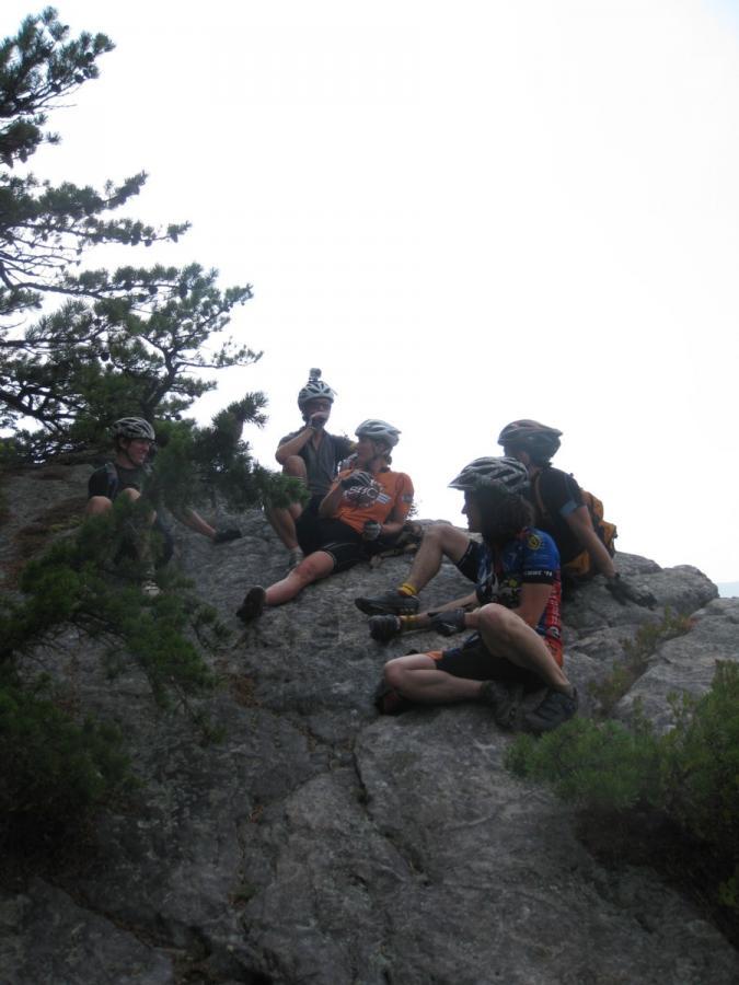 Five mountain bikers resting on a rocky outcrop in a natural outdoor setting, wearing helmets and cycling gear, surrounded by trees and a hazy sky. North Fork Mountain Trail mountain bike trail.