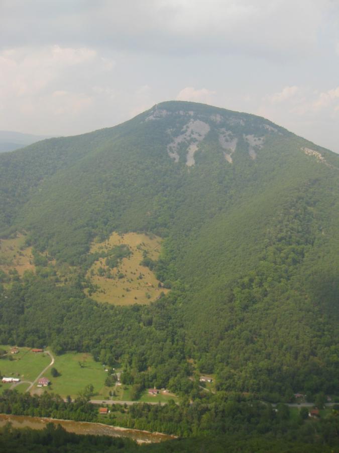 A panoramic view of a mountainous landscape with lush green hills, a prominent peak in the distance, and a valley below featuring patches of farmland and several scattered houses. The sky is partly cloudy, creating a serene and tranquil atmosphere. North Fork Mountain Trail mountain bike trail.