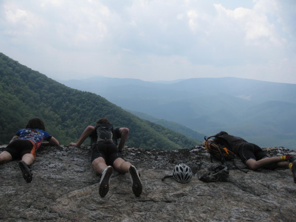 Three individuals lie on a rocky ledge, looking out over a vast mountainous landscape. The scene features lush green hills and rolling mountains under a cloudy sky. A bicycle helmet and gear are placed nearby on the rocky surface. North Fork Mountain Trail mountain bike trail.