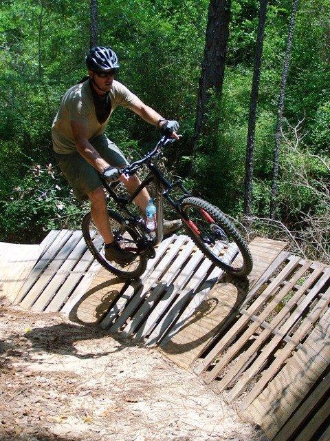 A mountain biker navigating a wooden ramp on a forest trail, wearing a helmet and sunglasses while performing a trick, with lush greenery in the background. Games Loop mountain bike trail.
