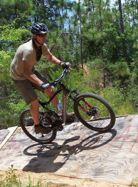 A man wearing a helmet and gloves rides a mountain bike up a wooden ramp in a forested area, surrounded by tall trees. He is focused and in motion, with his front wheel lifted off the ramp as he navigates the terrain. The ground is sandy and the ramp is marked with colorful graffiti. Games Loop mountain bike trail.