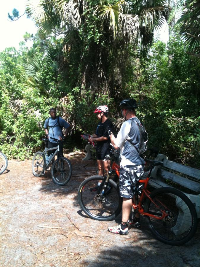 Three cyclists take a break on a forested trail. They are gathered around their mountain bikes. The surroundings are lush with green foliage and palm trees, creating a natural, sunny environment. One cyclist, wearing a red helmet, is gesturing as he speaks, while the others listen attentively. A bench is visible in the background. Okeeheelee Park / Pinehurst / Green Acres Freedom Park mountain bike trail.