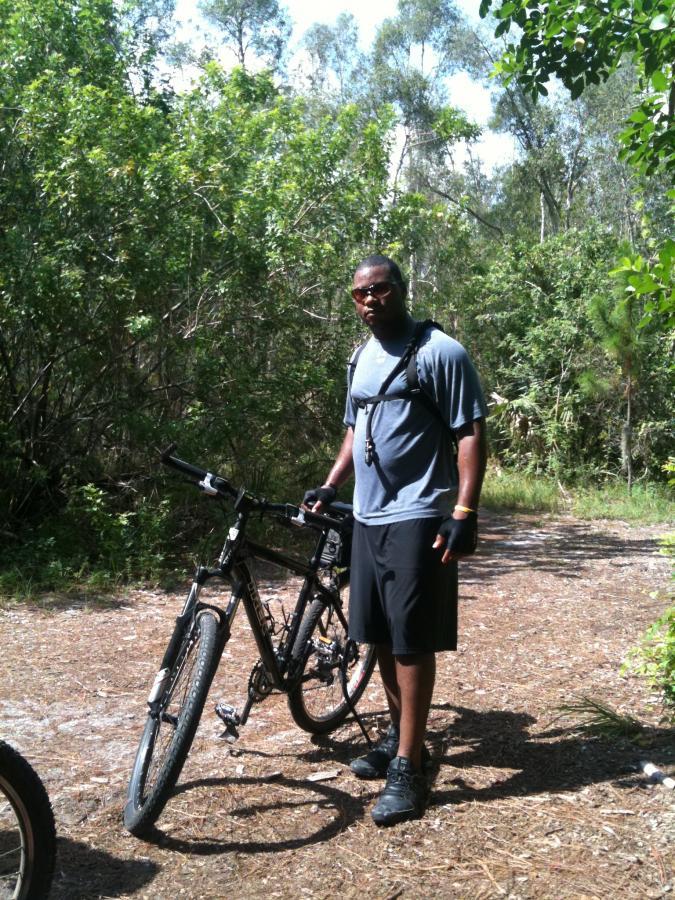 A person standing beside a mountain bike on a dirt path surrounded by greenery. The individual is wearing a gray athletic shirt and black shorts, with a bike helmet resting on their head and gloves on their hands. The background features trees and underbrush typical of a natural trail. Okeeheelee Park / Pinehurst / Green Acres Freedom Park mountain bike trail.