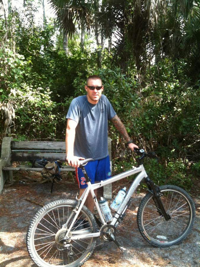 A man wearing sunglasses and athletic clothes stands beside his mountain bike in a wooded area. He is leaning on the bike, which has two water bottles mounted on the frame. In the background, there is a park bench and dense greenery typical of a natural setting. Okeeheelee Park / Pinehurst / Green Acres Freedom Park mountain bike trail.