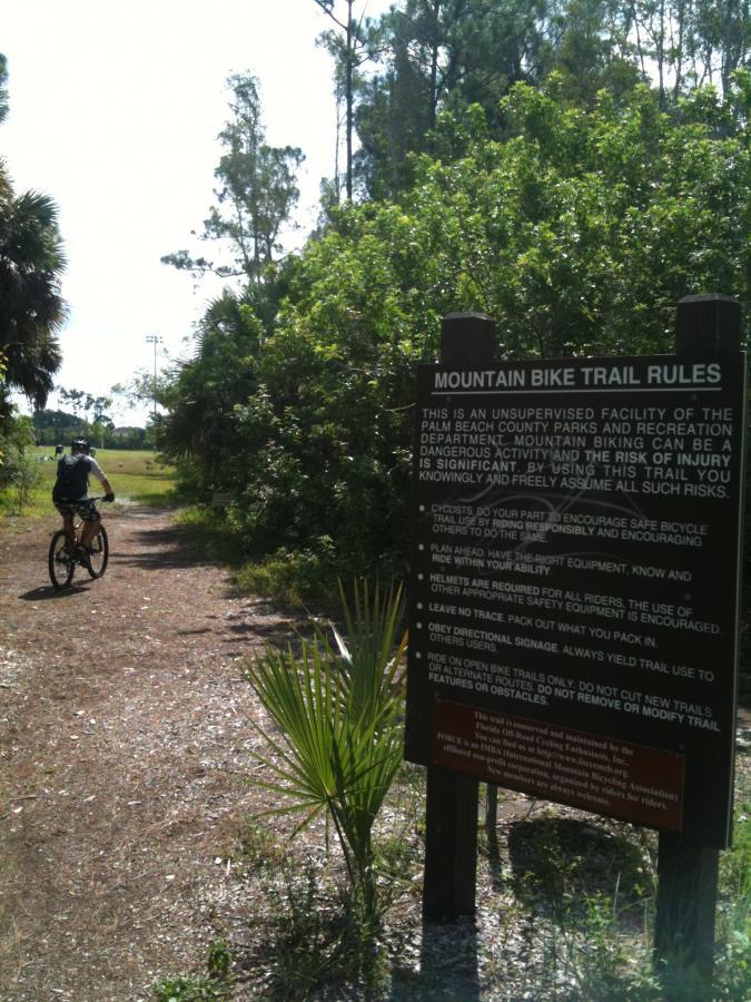 A mountain biker riding along a dirt trail surrounded by greenery, with a sign displaying the rules for mountain bike trails in a park. The sign emphasizes safety, helmet use, and encourages responsible biking behavior. Okeeheelee Park / Pinehurst / Green Acres Freedom Park mountain bike trail.