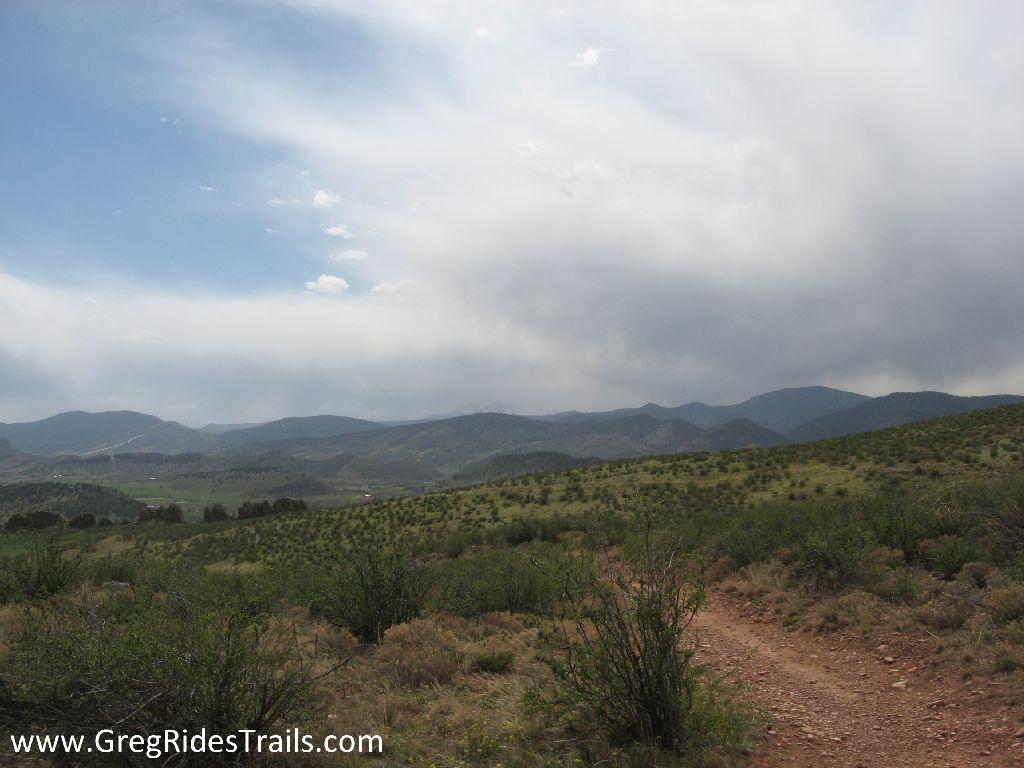 A scenic view of rolling hills and mountains under a cloudy sky, with a dirt path winding through grassy terrain and sparse vegetation. The landscape features a mix of shrubbery and open fields, capturing the natural beauty of the area. Devil's Backbone mountain bike trail.