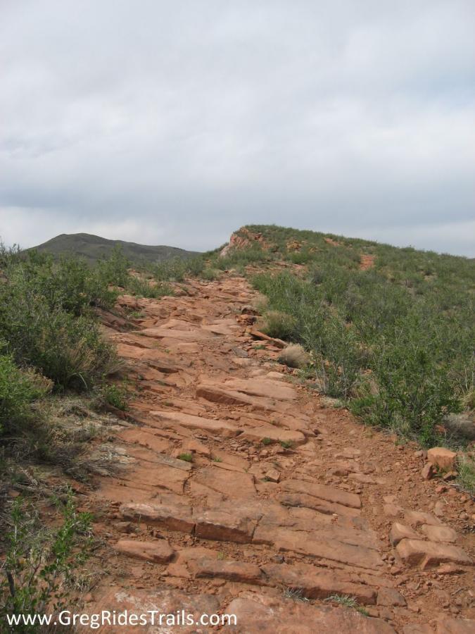 A rocky hiking trail winding upwards through sparse vegetation under a cloudy sky, with a hill visible in the background. Devil's Backbone mountain bike trail.
