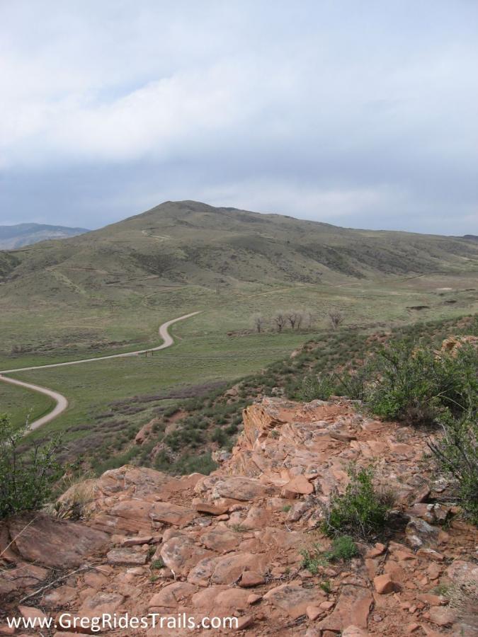 A scenic view of rolling green hills under a cloudy sky, featuring a winding dirt path. In the foreground, there are rocky terrain and sparse vegetation, with the hills fading into the background. Devil's Backbone mountain bike trail.