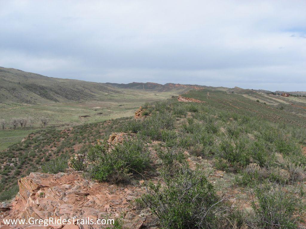 A scenic view of rolling hills and a rocky ridge under a partly cloudy sky, featuring patches of green grass and scattered bushes in the foreground. The landscape exhibits a mix of terrain with distant hills rising in the background. Devil's Backbone mountain bike trail.