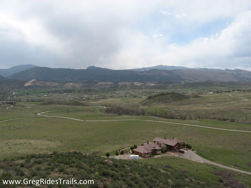 A scenic view of a rural landscape featuring a large house with a red tile roof situated on a green hillside, surrounded by rolling hills and distant mountains under a cloudy sky. The winding road leads up to the house, while patches of vegetation dot the landscape. Devil's Backbone mountain bike trail.