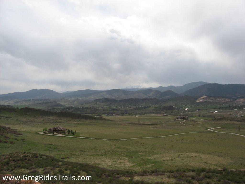 A panoramic view of rolling hills and mountains under a partly cloudy sky, featuring a grassy landscape with a winding road leading to a small house. The distant mountains are partially obscured by clouds, creating a serene and picturesque rural scene. Devil's Backbone mountain bike trail.
