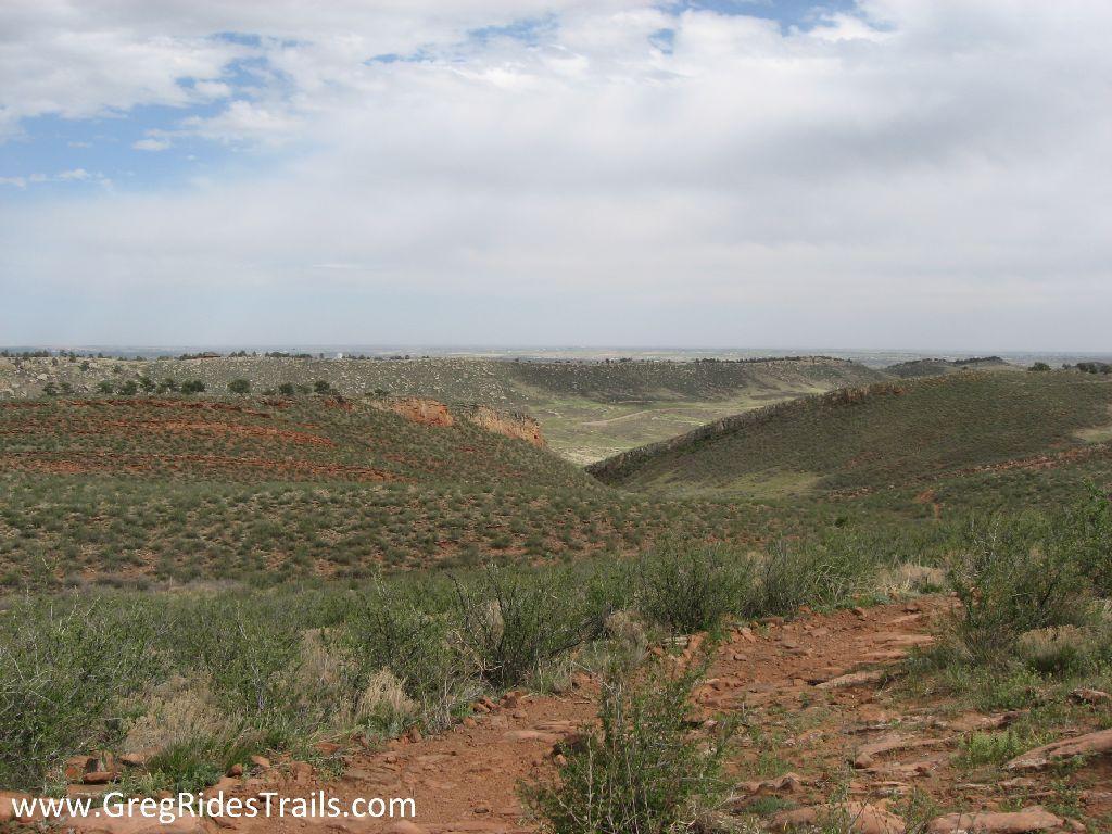 A panoramic view of rolling hills covered with green vegetation and patches of red earth under a cloudy sky. The landscape features a wide expanse of natural terrain, with shrubs and small plants scattered throughout the foreground and a distant horizon fading into the background. Devil's Backbone mountain bike trail.