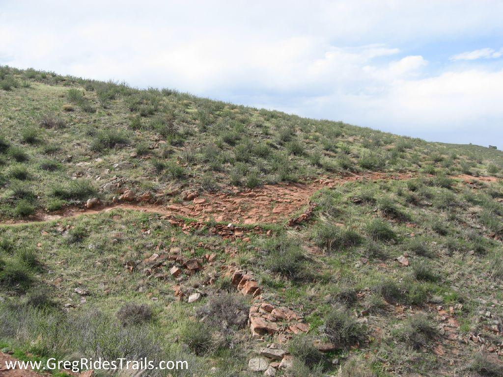A winding dirt trail curves along a grassy hillside, surrounded by small shrubs and rocks under a partly cloudy sky. The trail is set against a natural landscape, showcasing the terrain's rugged features. Devil's Backbone mountain bike trail.