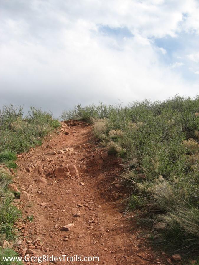 A rocky dirt trail winding upward through a grassy landscape under a partly cloudy sky. The trail features exposed red soil and scattered rocks, surrounded by shrubs and small plants on either side. Devil's Backbone mountain bike trail.