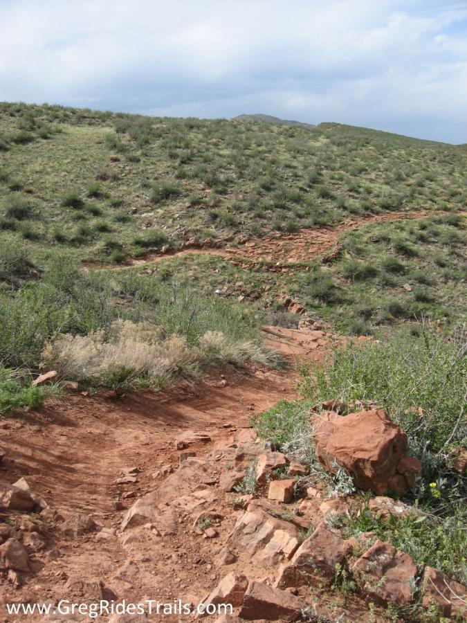 A winding dirt trail with a rocky path leads through grassy hills under a cloudy sky. The landscape features patches of green vegetation and scattered rocks along the trail. Devil's Backbone mountain bike trail.