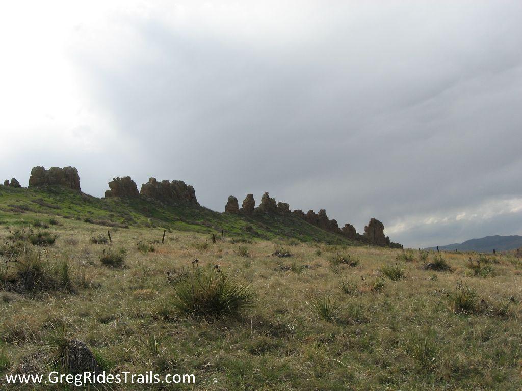 A wide view of a grassy landscape featuring rocky formations on a hillside under a cloudy sky. The foreground showcases sparse vegetation, while a line of jagged rock outcrops stretches across the horizon. Devil's Backbone mountain bike trail.