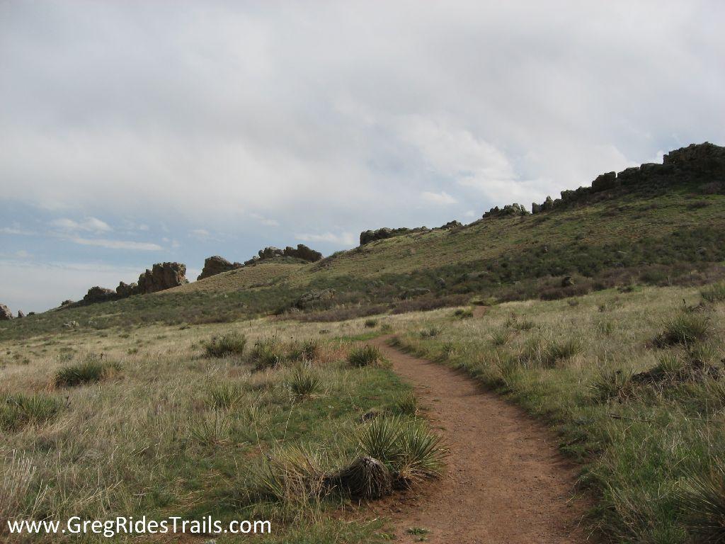 A scenic dirt path winding through grassy terrain, leading toward a rocky hillside under a cloudy sky. Devil's Backbone mountain bike trail.