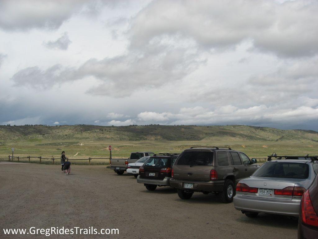 A gravel parking area with several parked cars, set against a backdrop of rolling hills and cloudy skies. Two individuals are seen in the foreground, one kneeling and the other standing near a sign. The landscape is green and expansive, suggesting an outdoor recreational area. Coyote Ridge mountain bike trail.