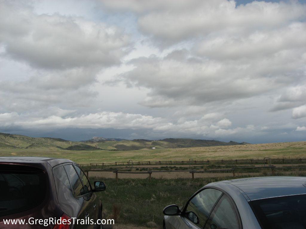 A scenic view of rolling green hills under a partly cloudy sky, with two cars parked in the foreground. The landscape features a wooden fence in the distance, adding to the serene rural setting. Coyote Ridge mountain bike trail.