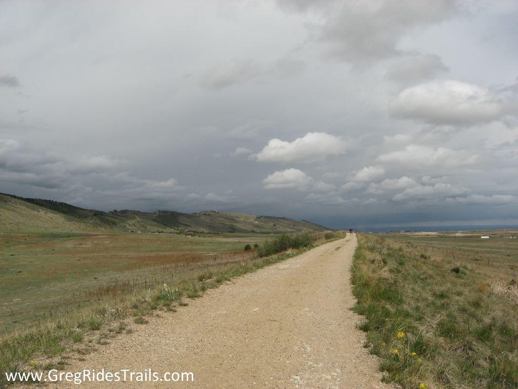 A wide, unpaved trail stretches along a grassy landscape, flanked by rolling hills under a cloudy sky. The path leads into the distance, suggesting a scenic journey through nature. Coyote Ridge mountain bike trail.