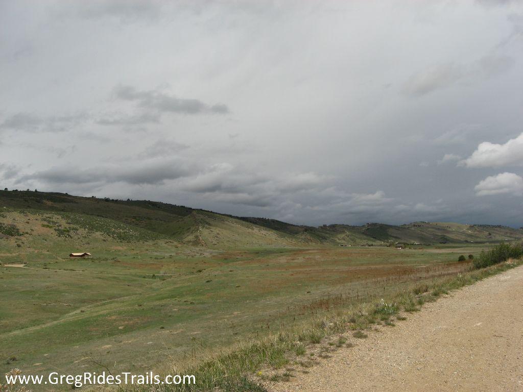A scenic view of rolling hills and an expansive grassy landscape under a cloudy sky, with a dirt path leading through the foreground. A small wooden structure is visible in the distance, surrounded by open terrain. Coyote Ridge mountain bike trail.