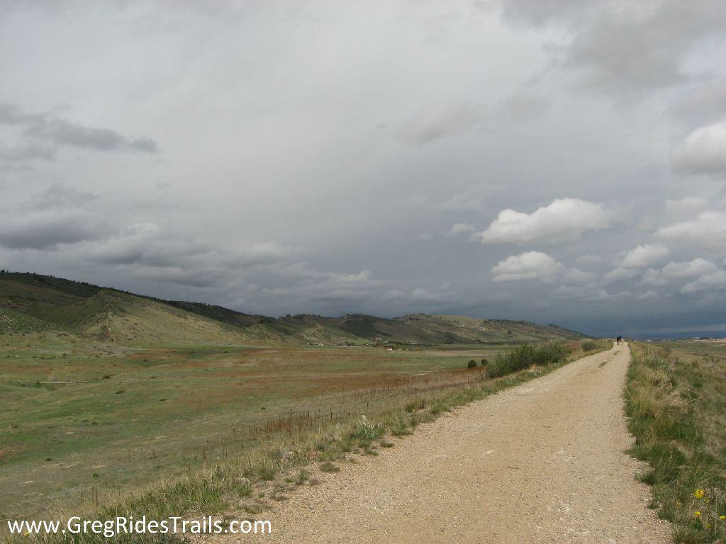 A wide dirt trail runs through an expansive landscape, flanked by rolling hills and patches of green grass. The sky is overcast with gray clouds, suggesting an impending storm. In the distance, a few figures can be seen walking along the path. The scene captures a serene yet dramatic natural environment. Coyote Ridge mountain bike trail.
