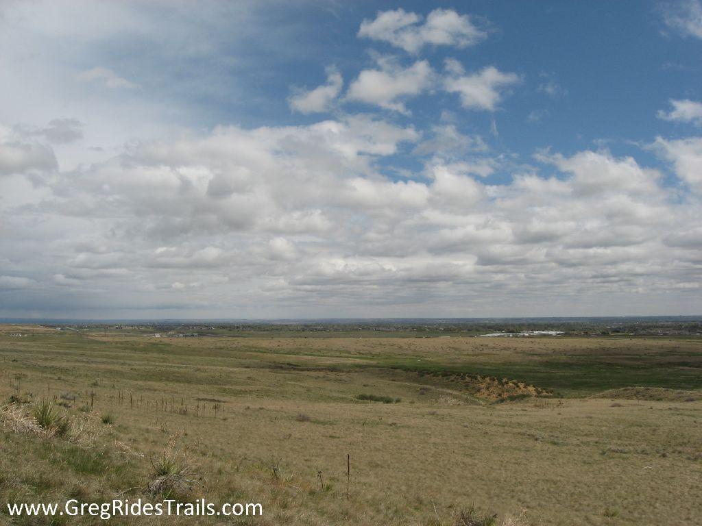 Landscape view of open grassland under a cloudy sky, showcasing rolling plains and distant hills. The foreground features sparse vegetation, while the horizon includes scattered buildings and a blue sky filled with white clouds. Coyote Ridge mountain bike trail.