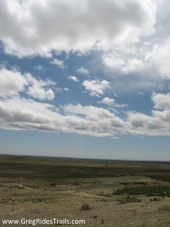 A panoramic view of a landscape under a blue sky filled with fluffy white clouds, featuring rolling hills and green fields in the foreground. A winding trail can be seen cutting through the terrain, leading into the distance. Coyote Ridge mountain bike trail.