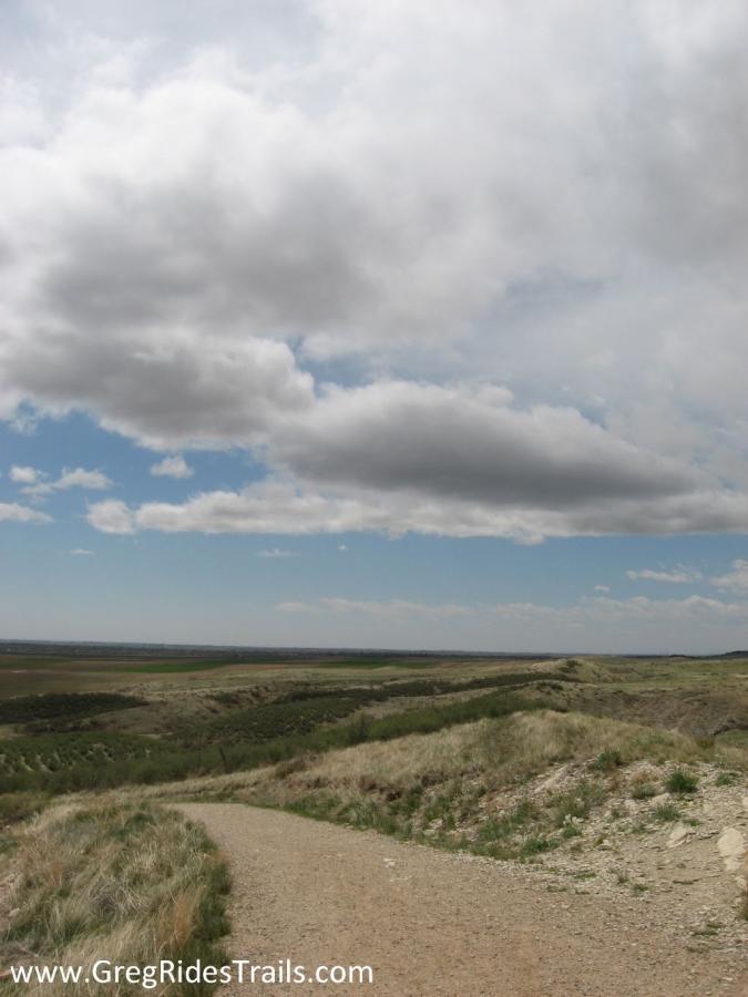 A scenic view of a winding dirt path leading through grassy hills under a partly cloudy sky. The landscape suggests a tranquil outdoor setting, with distant horizons and patches of green vegetation. Coyote Ridge mountain bike trail.