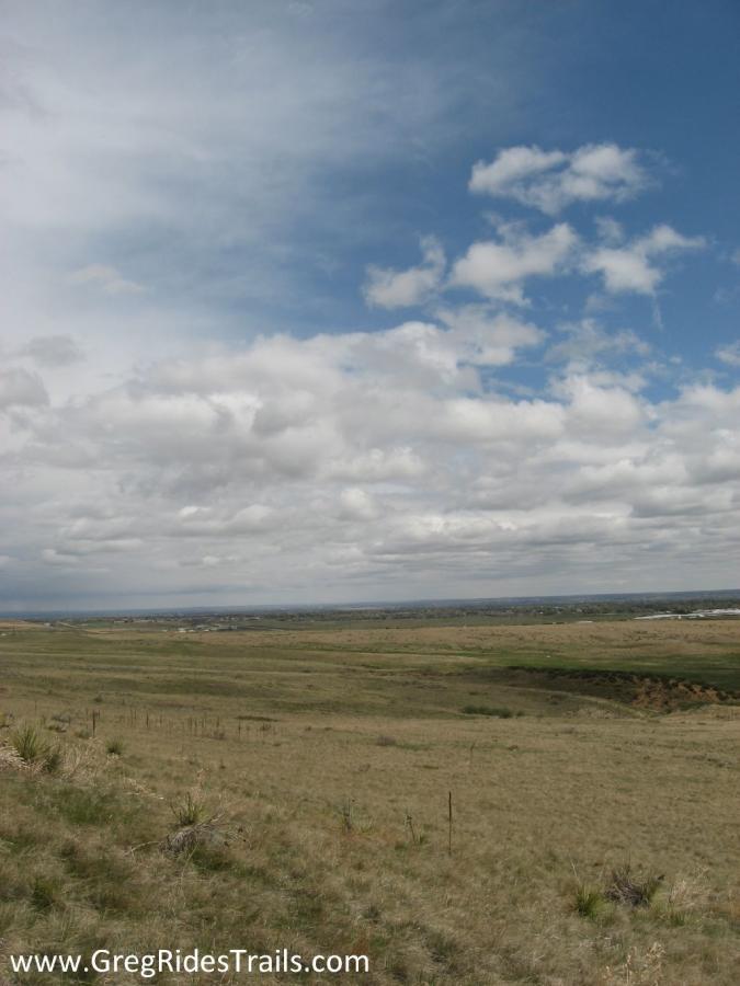 A scenic landscape featuring expansive grassy plains under a dynamic sky filled with clouds, with hints of distant hills and structures in the background. The image captures a serene and tranquil natural setting. Coyote Ridge mountain bike trail.