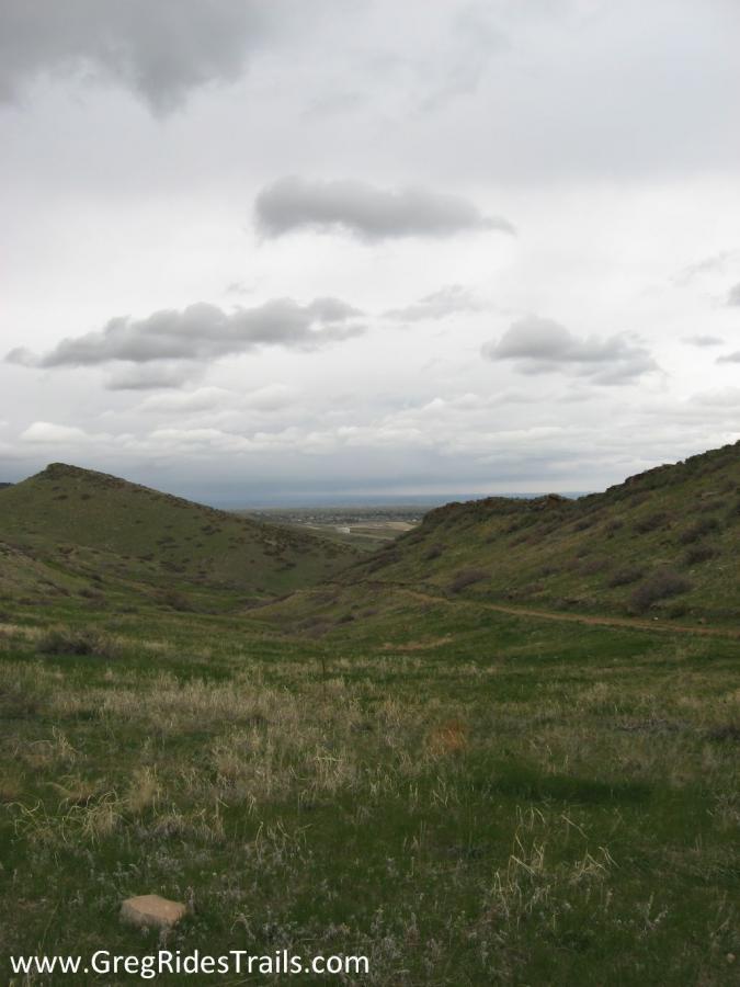 A panoramic view of rolling green hills under a cloudy sky, showcasing a natural landscape with a winding trail and patches of grass. The valley below extends into the distance, creating a serene and picturesque outdoor setting. Coyote Ridge mountain bike trail.