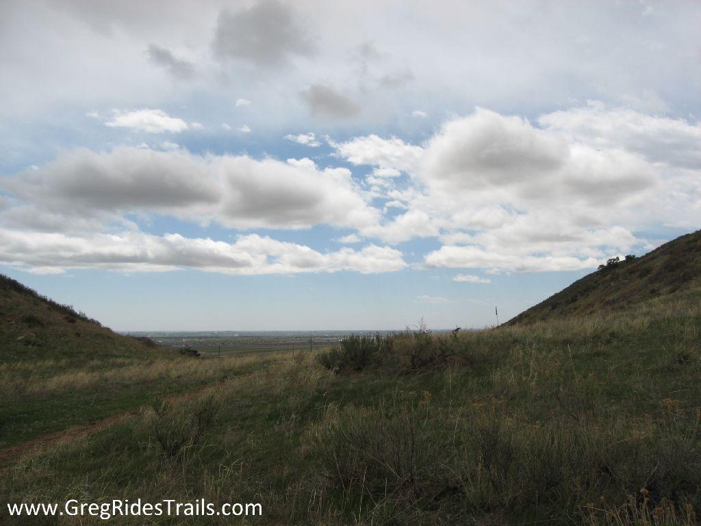 A scenic view of rolling hills with green grass under a partly cloudy sky. The horizon is visible in the distance, with gentle slopes rising on either side. The landscape conveys a sense of tranquility and natural beauty. Coyote Ridge mountain bike trail.