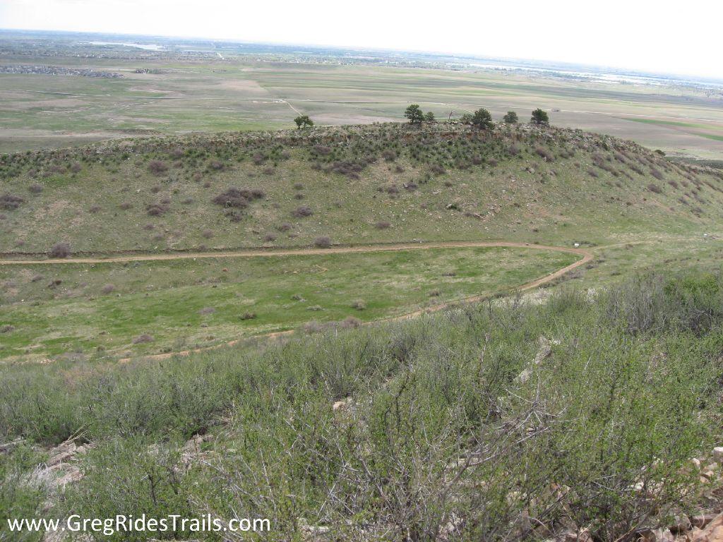 A scenic overlook of a winding dirt trail along a hillside, surrounded by lush green grass and scattered shrubs. In the distance, a flat landscape stretches out toward a body of water under a cloudy sky. Coyote Ridge mountain bike trail.