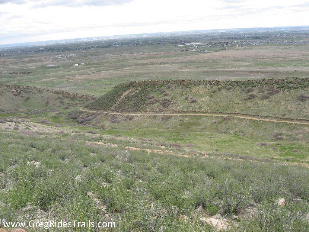 A panoramic view of rolling hills and green landscapes under a cloudy sky. The foreground features a grassy slope dotted with small shrubs, while a winding dirt trail leads through the hills in the background. The expansive scenery includes patches of cultivated land and distant structures, showcasing a blend of natural and rural environments. Coyote Ridge mountain bike trail.