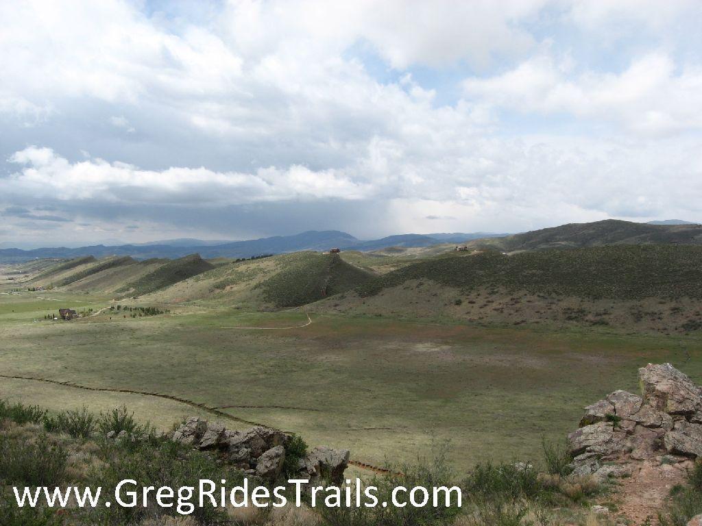 A scenic view of rolling hills and open fields under a cloudy sky. The landscape features gentle slopes and patches of greenery, with a hint of rugged terrain in the foreground. A dirt path winds through the fields, leading toward the distant mountains on the horizon. Coyote Ridge mountain bike trail.