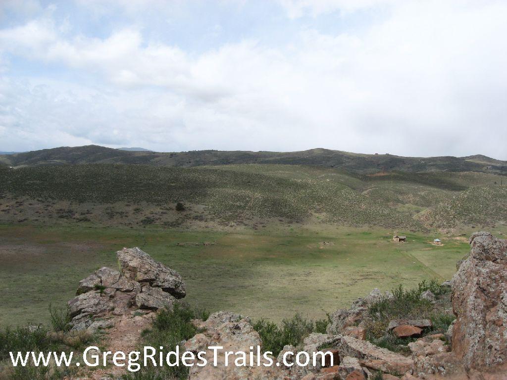 A wide landscape view showing rolling hills with green vegetation under a partly cloudy sky. In the foreground, there are various rock formations and scattered bushes. In the distance, a small structure is visible among the hills, suggesting a rural or natural setting. Coyote Ridge mountain bike trail.