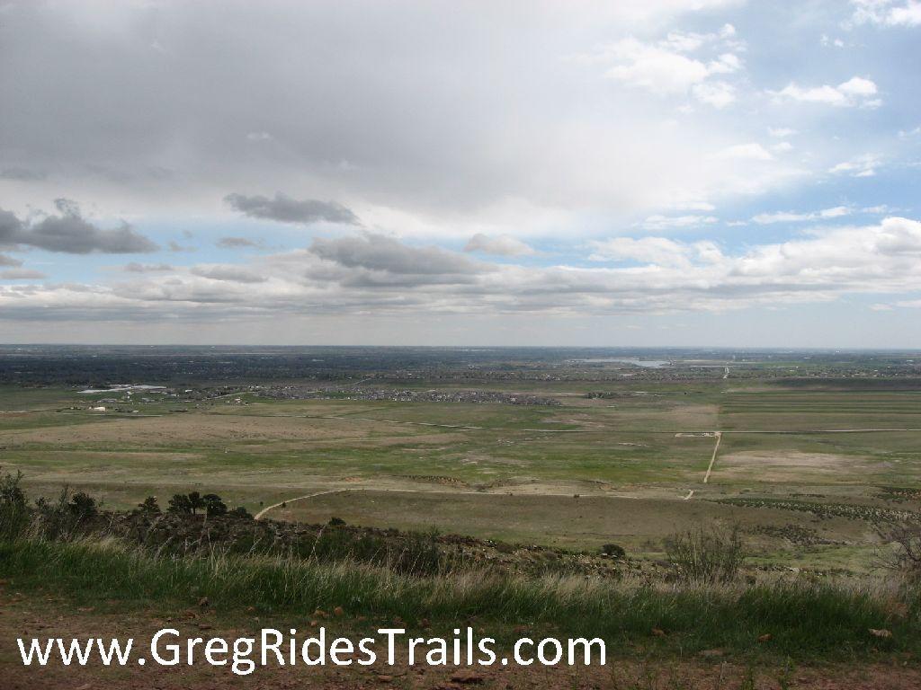 A panoramic view of an expansive landscape featuring green fields, patches of water, and distant urban structures under a cloudy sky. The foreground includes grasses and a slight elevation, making it a scenic overlook. Coyote Ridge mountain bike trail.