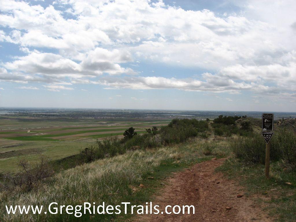 A scenic view from a hillside trail overlooking a vast landscape of fields and valleys, with a partly cloudy sky above. A trail marker stands to the right, indicating the pathway for hikers. Coyote Ridge mountain bike trail.
