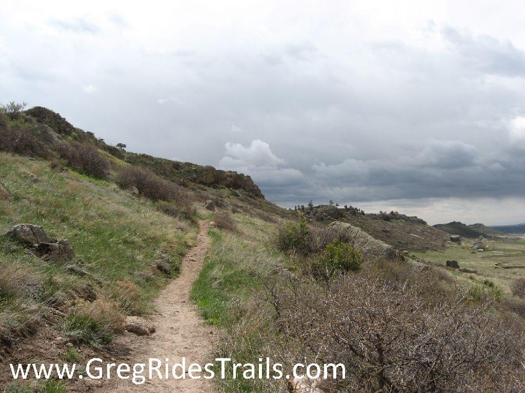 A winding dirt trail leads through lush green grass and shrubs, flanked by rocky outcrops under a cloudy sky. The peaceful landscape suggests a serene hiking environment in a natural setting. Coyote Ridge mountain bike trail.