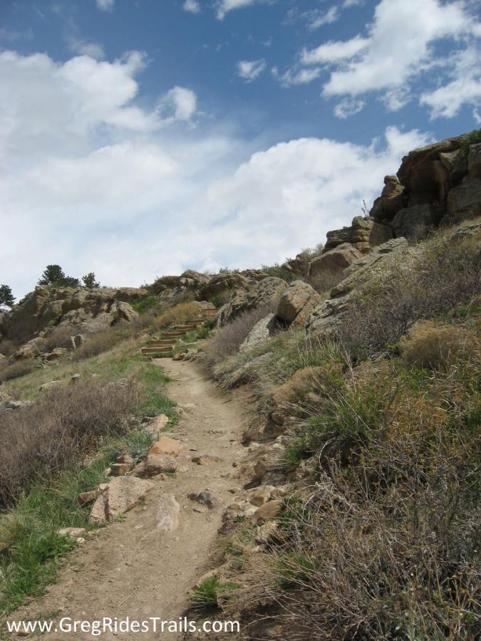 A winding dirt trail leads upward through rocky terrain and sparse vegetation, under a partly cloudy sky. Steps can be seen ascending along the path, surrounded by grass and shrubs. Coyote Ridge mountain bike trail.