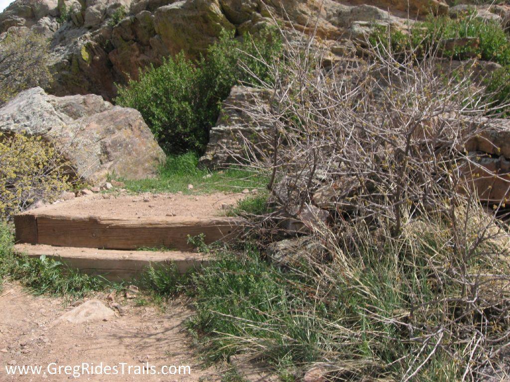 A small wooden step leads up a dirt path, surrounded by rocky outcrops and sparse vegetation, including dry bushes and patches of grass. The scene showcases a natural outdoor setting in a rugged terrain. Coyote Ridge mountain bike trail.