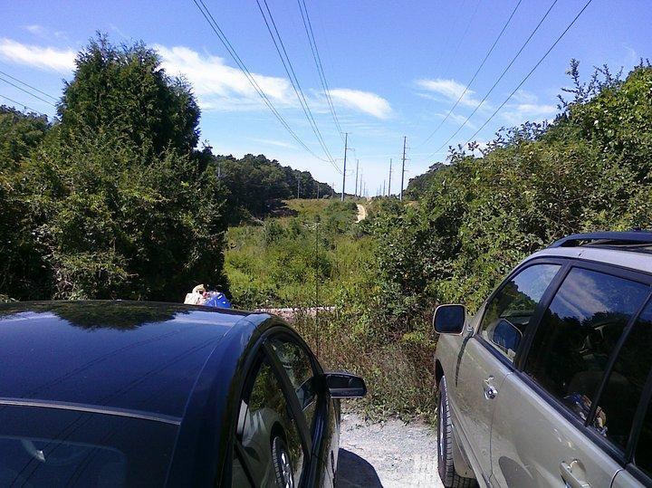 Two parked cars on a gravel road with lush greenery on either side. In the background, power lines stretch across a clear blue sky, and a dirt path leads into the wooded area. The scene is bright and sunny, indicating a pleasant day in a natural setting. West Barnstable Conservation Area mountain bike trail.