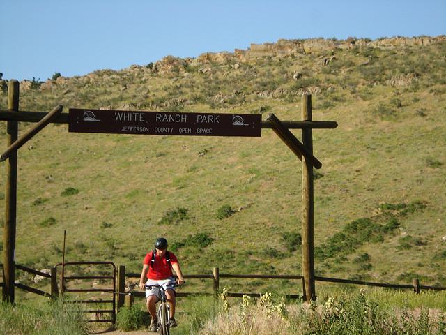A cyclist riding along a trail in front of the entrance sign to White Ranch Park, a grassy area with rolling hills and sparse vegetation in the background. The sign indicates that the park is part of Jefferson County Open Space. White Ranch mountain bike trail.