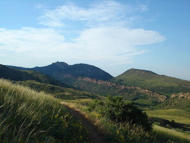 A scenic view of rolling green hills under a blue sky, with a dirt path winding through tall grass in the foreground and rocky mountain peaks in the background. White Ranch mountain bike trail.
