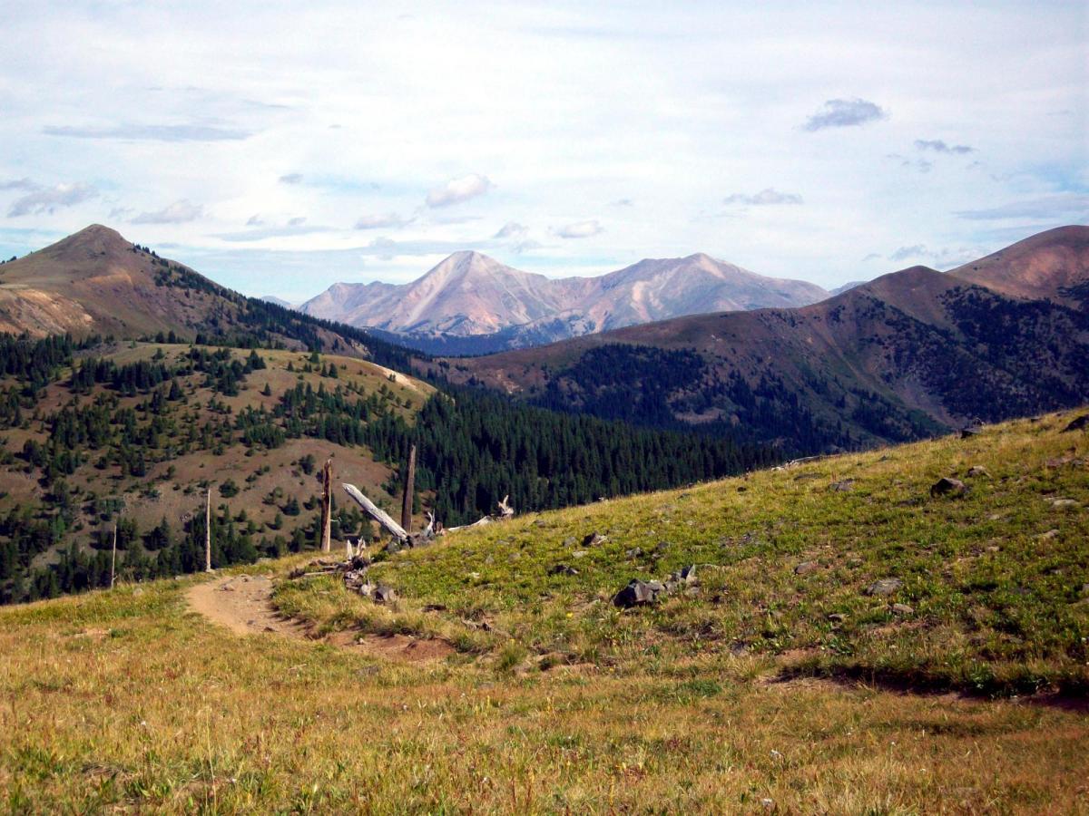 A scenic view of rolling hills and mountains under a partly cloudy sky, showcasing green grass and patches of trees in the foreground. The background features tall mountains with varying shades of brown and gray, highlighting the natural beauty of the landscape. Monarch Crest Trail mountain bike trail.